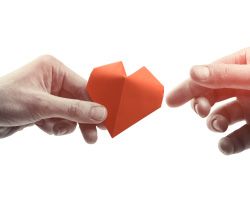 A hand reaching out to take a paper heart from another hand.