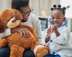 A woman holding a large teddy bear laughs with a young girl in a doctor costume.